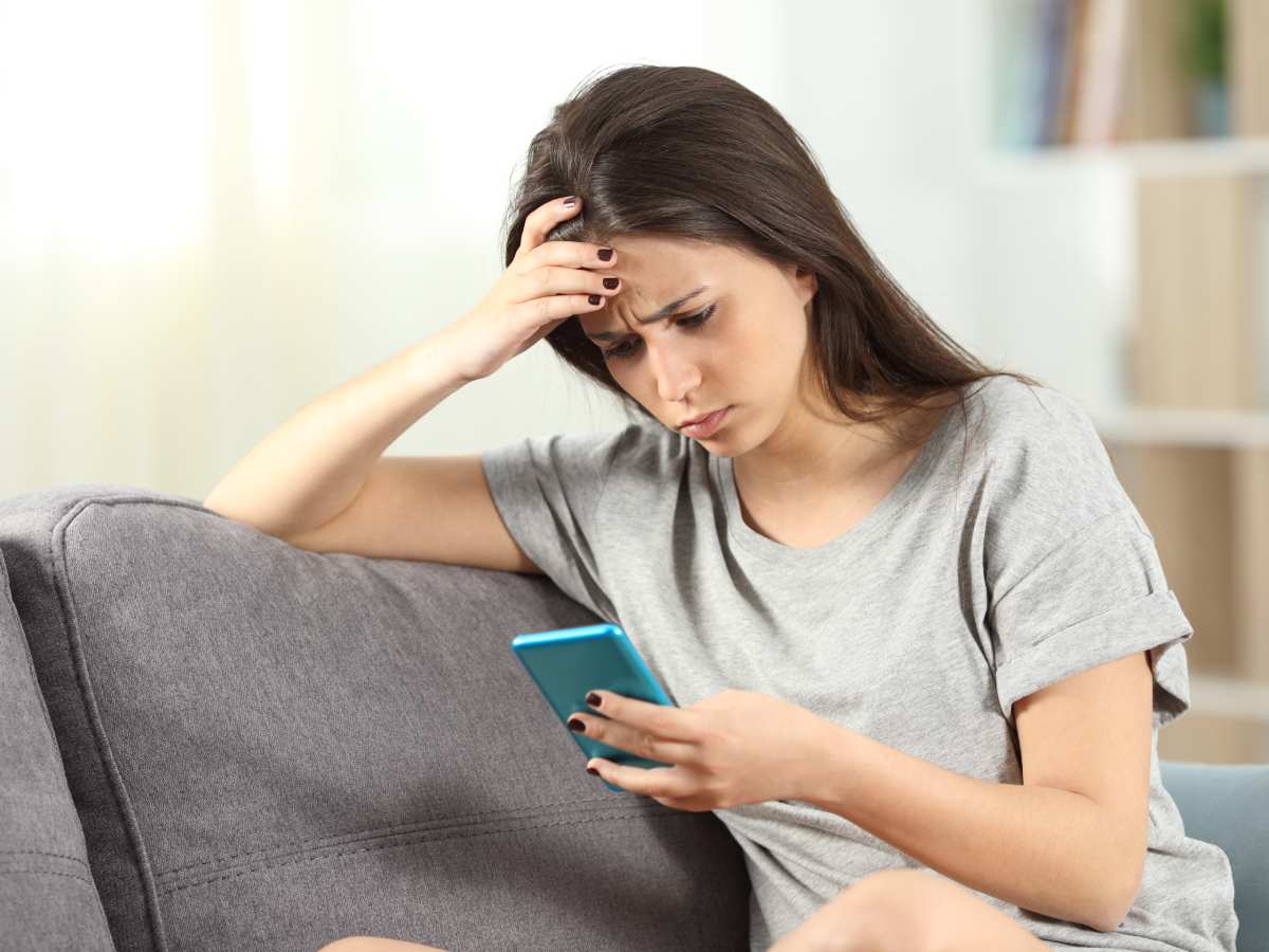 Zabrinuta tinejdžerka sedi na kauču i gleda u ekran mobilnog telefona